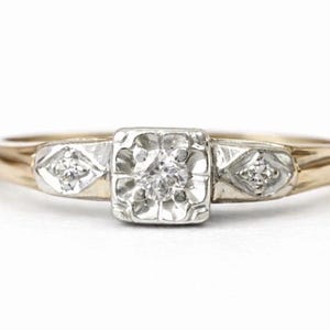 May include: A gold ring featuring a central square-shaped setting with a single round diamond, flanked by two smaller diamond-set accents in diamond-shaped white metal mounts. The band is crafted from polished yellow gold with a smooth finish.