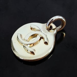 Puede incluir: Colgante dorado con un logotipo de doble C sobre una base ovalada. El colgante tiene un lazo en la parte superior para una cadena. El logotipo es de un oro más claro que la base. El fondo es negro.