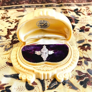Puede incluir: Un anillo de diamantes con una piedra central de talla marquesa y dos piedras laterales, engastado en una banda plateada, presentado en un joyero crema abierto. El joyero tiene un forro de terciopelo morado oscuro. El joyero tiene el texto "THE SIGN OF QUALITY".