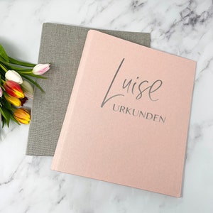 Könnte beinhalten: Ein rosa-grauer, stoffbezogener Ordner mit dem Text "Luise URKUNDEN" auf der Vorderseite.