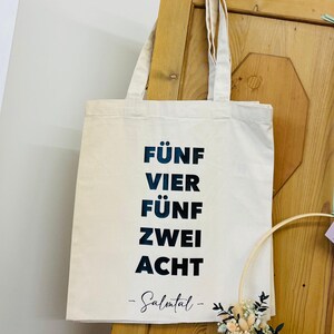 Könnte beinhalten: Eine weiße Stofftasche mit schwarzem Text, der "Fünf Vier Fünf Zwei Acht -Salmtal-" lautet.