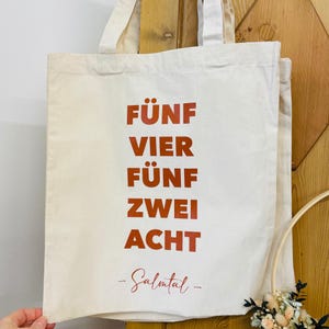 Könnte beinhalten: Eine weiße Stofftasche mit dem Aufdruck "FÜNF VIER FÜNF ZWEI ACHT -Salmtal-" in orange.