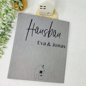 Könnte beinhalten: Grauer Leinenbezogener Album mit dem Text "Hausbau Eva & Jonas" und einer kleinen Haus-Illustration mit der Nummer 16 an der Tür.