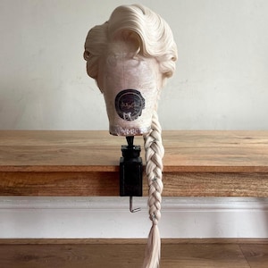 Könnte beinhalten: Hellblonde Perücke mit einem langen, geflochtenen Pferdeschwanz, auf einem Schaufensterkopf frisiert. Die Perücke hat voluminöse Locken am Oberkopf und ist mit einer schwarzen Klemme auf einer Holzoberfläche befestigt. Silberne Akzente sind in den Zopf eingewebt.