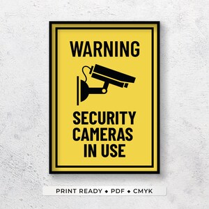 May include: A yellow warning sign with black text and a black silhouette of a security camera. The sign reads "WARNING SECURITY CAMERAS IN USE".