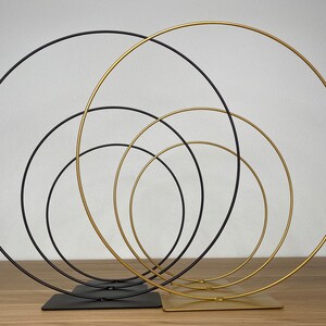 May include: Set of five interlocking metal hoops, with three gold hoops and two black hoops. The hoops are arranged in a circular pattern, with the largest hoop on the outside and the smallest hoop on the inside. The hoops are standing on a wooden surface.