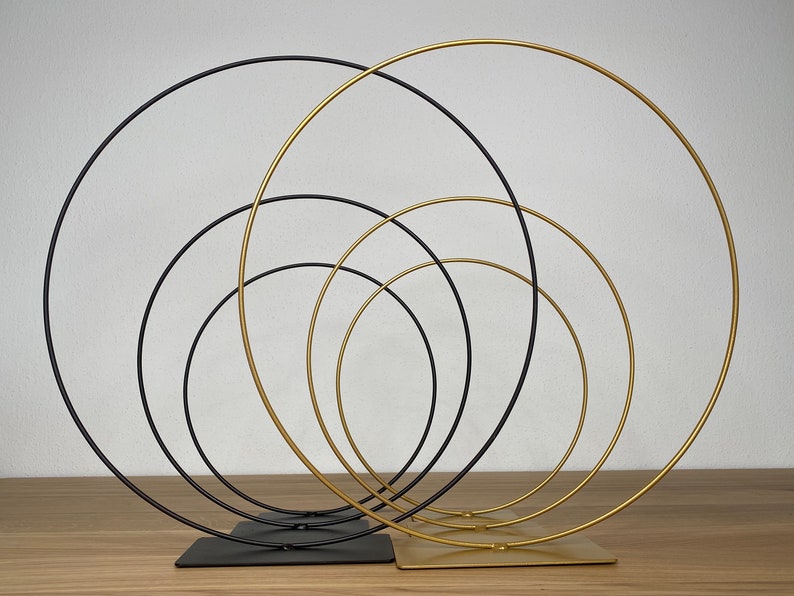 May include: Set of five interlocking metal hoops, with three gold hoops and two black hoops. The hoops are arranged in a circular pattern, with the largest hoop on the outside and the smallest hoop on the inside. The hoops are standing on a wooden surface.