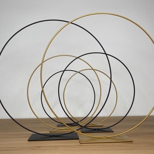 May include: A set of five metal hoops, four black and one gold, arranged in a concentric circle pattern. The hoops are on stands and are ideal for displaying flowers or other decorative items.