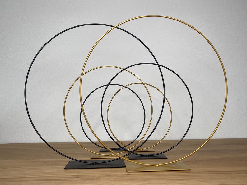 May include: A set of five metal hoops, four black and one gold, arranged in a concentric circle pattern. The hoops are on stands and are ideal for displaying flowers or other decorative items.