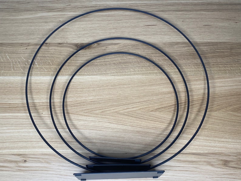 May include: Three black metal hoops of different sizes stacked on top of each other. The hoops are connected by a flat black metal base.