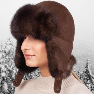 Puede incluir: Un gorro trapero marrón con orejeras de piel. El gorro está hecho de una tela suave de color marrón y tiene un ala ancha. El ribete de piel es de un color marrón oscuro intenso y es muy suave.