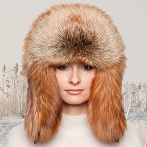 Peut inclure: Un chapeau en fourrure marron et roux avec des cache-oreilles. Le chapeau a un aspect doux et texturé et couvre la tête et les oreilles. L'arrière-plan montre un paysage hivernal avec de la neige et des herbes hautes.