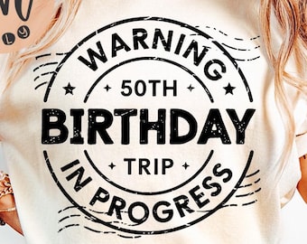 May include: Cream-colored t-shirt with a black circular graphic that reads "WARNING 50TH BIRTHDAY IN PROGRESS." The design includes stars and the text "TRIP." The shirt is paired with blue jeans.