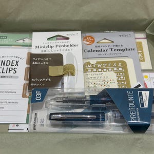 Puede incluir: Un set de papelería en una caja de cartón que contiene una pluma estilográfica azul Platinum Prefounte, un portalápices miniclip de metal dorado, un paquete de clips de índice y una plantilla de calendario, todo en su embalaje original.