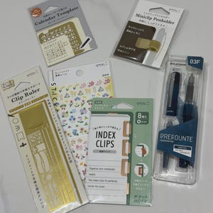 Könnte beinhalten: Eine Sammlung von Schreibwaren, bestehend aus einer goldenen Kalendervorlage, einem goldenen Miniclip-Stiftehalter, einem goldenen Clip-Lineal, einem Bogen Blumenaufkleber, einer Packung mit acht Index-Clips und einem blauen Platinum Prefounte Füllfederhalter.