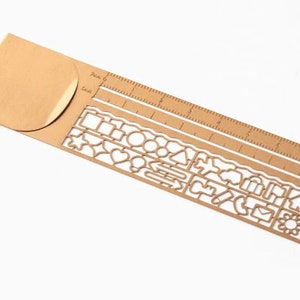 Può includere: Un righello in metallo color oro con un segnalibro di carta marrone attaccato. Il righello ha una scala standard in pollici e una varietà di forme ritagliate lungo il bordo inferiore, tra cui stelle, cuori, cerchi e case.