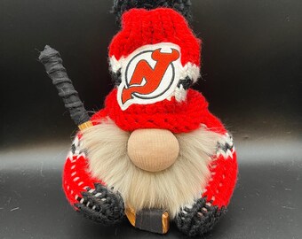 May include: A red and black gnome wearing a knitted hat with the New Jersey Devils logo. The gnome is holding a hockey stick.