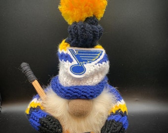 May include: A blue and yellow gnome wearing a crocheted hat with a St. Louis Blues logo and holding a hockey stick.