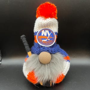 Op de afbeelding: Een gebreide gnome die een blauwe en oranje muts draagt met het logo van de New York Islanders. De gnome houdt een hockeystick vast.