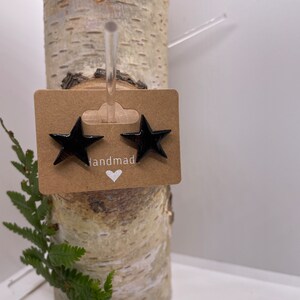May include: A pair of black star earrings on a birch log. The earrings are glossy and attached to a small brown tag with the word "handmade" and a heart symbol. Green fern fronds are in the foreground.