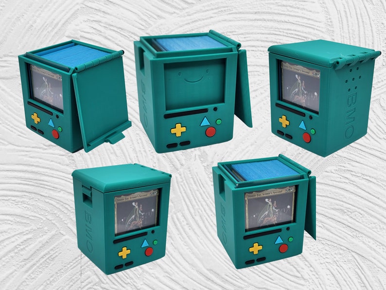 May include: A teal blue card game storage box with a hinged lid and a design inspired by the character BMO from the animated series Adventure Time. The box has a screen-like front panel with a card visible inside, and a set of buttons on the bottom. The text "BMO" is printed on the side of the box.