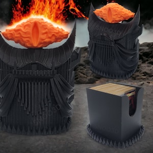 Può includere: Portacarte nero con un design stilizzato, con un elemento arancione a forma di occhio sopra. Il supporto ha una superficie strutturata e il testo "SNOO3D" è visibile. Il design evoca un tema fantasy.
