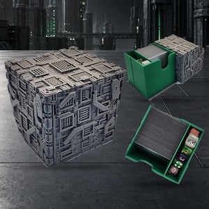 May include: A grey cube-shaped storage box with a complex mechanical surface texture. The box opens to reveal a green interior compartment containing a stack of rectangular cards and a row of five multi-coloured polyhedral dice.