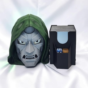 Op de afbeelding: Een 3D-geprint grijs masker met een groene capuchon naast een zwarte kaartenbak. De doos heeft een voorvak met daarin een blauwe dobbelsteen met witte stippen en een gouden dobbelsteen met het cijfer vier in het zwart gedrukt.