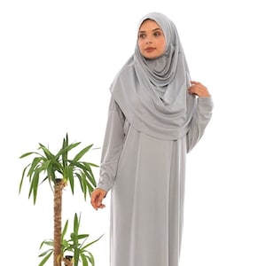 Könnte beinhalten: Ein hellgraues, langärmliges Kleid mit passendem Hijab. Das Kleid hat eine lockere Passform und ein schlichtes Design.