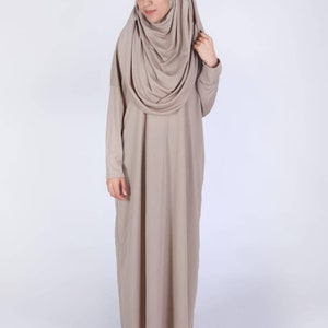 May include: A long-sleeved, floor-length dress and matching head covering in a neutral beige color. The dress has a simple, flowing design, and the head covering drapes elegantly around the shoulders.