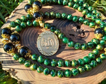 Può includere: Una collana di perline verdi e nere con un ciondolo dorato che presenta la parola araba per Dio, "Allah".