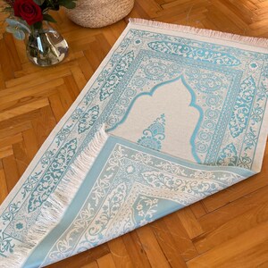 May include: A light blue and cream-colored prayer rug with an ornate floral design and fringed edges. The rug is partially folded, revealing the intricate patterns. The rug is placed on a wooden floor.