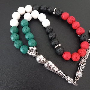 May include: A green, white and red prayer bead necklace with a silver tassel and a silver charm. The beads are made of lava rock and are strung on a silver chain.