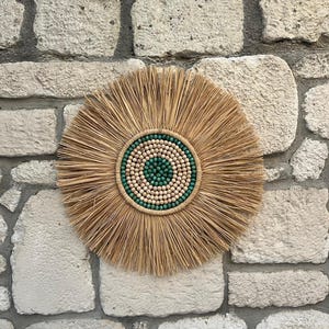 Puede incluir: Un adorno de pared redondo con un diseño de paja tejida natural. El centro presenta un patrón circular de pequeñas cuentas de madera de color claro y cuentas de color turquesa. La decoración de la pared está montada en una pared de piedra texturizada.