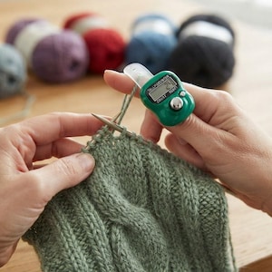 Può includere: Un contatore digitale verde con cinturino in plastica trasparente indossato su un dito, posizionato accanto a un lavoro a maglia verde salvia con motivo a trecce su ferri in metallo, con diversi gomitoli colorati sullo sfondo.