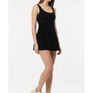 May include: A black athletic dress with a scoop neckline and a flared skirt. The dress is sleeveless and worn with cream-colored athletic shoes. The model is standing in front of a white background.