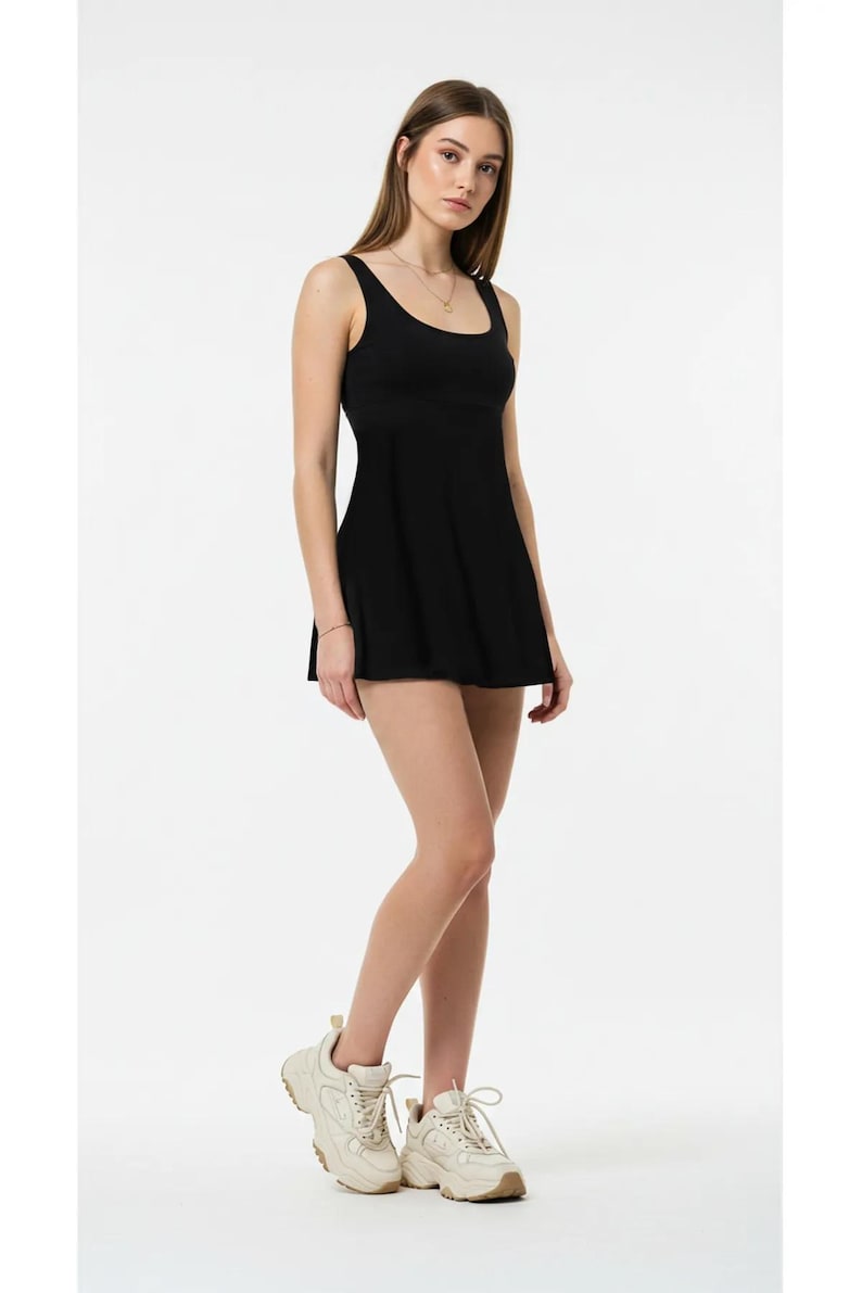 May include: A black athletic dress with a scoop neckline and a flared skirt. The dress is sleeveless and worn with cream-colored athletic shoes. The model is standing in front of a white background.
