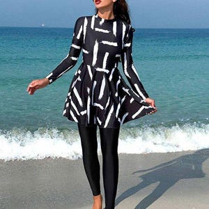 May include: A black and white long-sleeved swimsuit featuring a flared tunic top with a white brushstroke pattern and solid black leggings. The garment has a high neckline and provides full coverage, extending to the wrists and ankles.