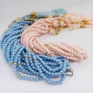 May include: An assortment of beaded necklaces in pastel colors. The necklaces are in shades of light blue and pink, with gold-colored accents and tassels. The beads appear to be round and uniform in size, creating a delicate and elegant look.