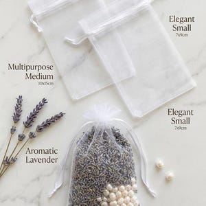 Op de afbeelding: Drie transparante witte organza zakjes met trekkoord. Twee lege zakjes zijn gelabeld Elegant Small 7x9cm en Multipurpose Medium 10x15cm. Eén gevuld zakje bevat gedroogde lavendel en witte parels, gelabeld Pearl & Jewelry Size 10x15cm.