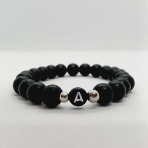 Könnte beinhalten: Schwarzes Perlenarmband mit dem weißen Buchstaben "A" auf einer schwarzen Perle. Das Armband besteht aus mattschwarzen Perlen und silberfarbenen Akzentperlen. Ein schlichtes, elegantes Accessoire.