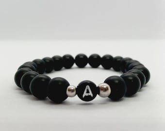 May include: Black beaded bracelet with the letter "A" in white on a black bead. The bracelet is made of matte black beads and silver-colored accent beads. The bracelet is a simple, elegant accessory.