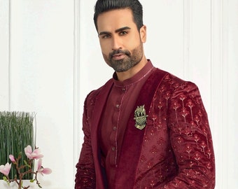 May include: A maroon velvet suit with a patterned design. The suit includes a long jacket with a mandarin collar and matching trousers. The jacket features a gold brooch with a floral design.