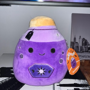May include: A plush purple potion bottle with a smiling face, black eyes, and a beige top. The bottle has a decorative emblem and a black tag. A tag reads "Squishmallows Kensington".