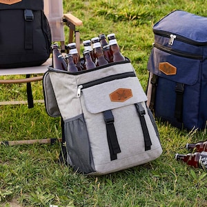 May include: Three insulated backpacks, two black and one gray, with a personalized name tag on the black backpack that says "AIE". The gray backpack has a zippered pocket and is filled with bottles of beer. The backpacks are on a wooden folding chair and grass.