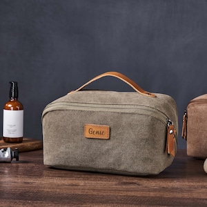 May include: A green canvas wash bag with a brown leather handle and zip. The bag has a brown leather patch with the word "Genie" written on it.