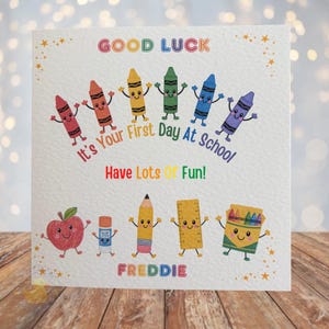May include: A square greeting card with a white textured background featuring colourful cartoon illustrations of crayons, an apple, a glue stick, a pencil, a ruler, and a crayon box. Text reads: GOOD LUCK It's Your First Day At School Have Lots Of Fun! FREDDIE