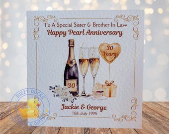 Può includere: Un biglietto bianco con dettagli dorati e un'illustrazione ad acquerello di bicchieri di champagne, una bottiglia di champagne, un cupcake e una scatola regalo. Il biglietto dice "To A Special Sister & Brother In Law Happy Pearl Anniversary" e "Jackie & George 16th July 1995".