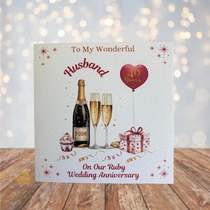 May include: A square anniversary card with a watercolour design. It features a champagne bottle, two filled glasses, a cupcake, and a gift box. Text includes "To My Wonderful Husband" and "On Our Ruby Wedding Anniversary". A red heart-shaped balloon.