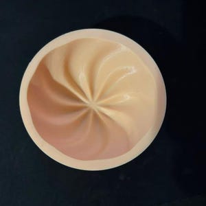 May include: A circular, light peach-colored silicone mold featuring a central, swirling, star-like pattern with radiating grooves. The mold has a smooth, rounded rim and a concave interior, designed for creating detailed, shaped items.
